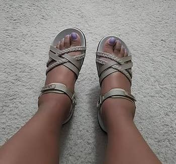 Person wearing strappy sandals with braided details, showing lavender-painted toenails connected a carpeted floor
