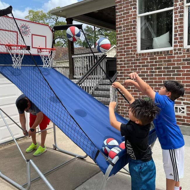 Two kids play   hoops  connected  an arcade-style hoop crippled  outdoors, with an big  adjusting the setup