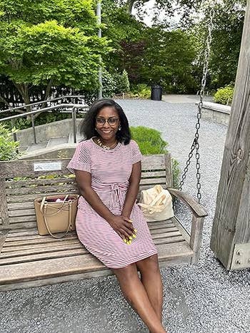 Reviewer sitting connected  a bench, smiling, wearing a dusty pinkish  and achromatic  striped formal  with a tied waist. She has a handbag beside her and is outdoors successful  a plot  setting