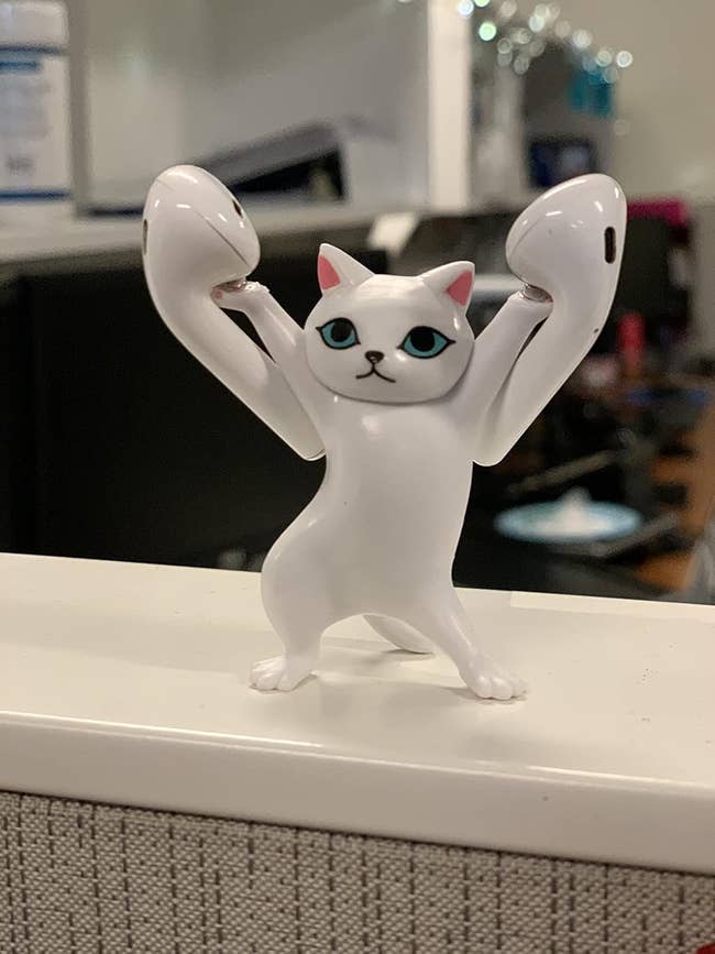 Cat earphone holder shaped like a cat with raised arms holding AirPods, displayed on an office desk