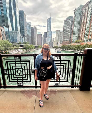 Person in a casual summer outfit with sunglasses stands on a riverside walkway with city skyscrapers in the background
