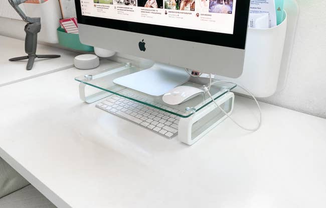 A clear glass monitor stand on a desk with an iMac, keyboard, mouse, and organizer with stationery items