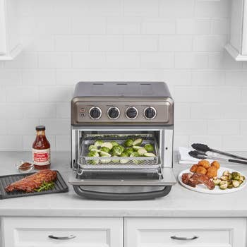 Countertop oven with 4 dials and a tray of roasted Brussels sprouts. Surrounding plates person ribs, baked bites, and much Brussels sprouts