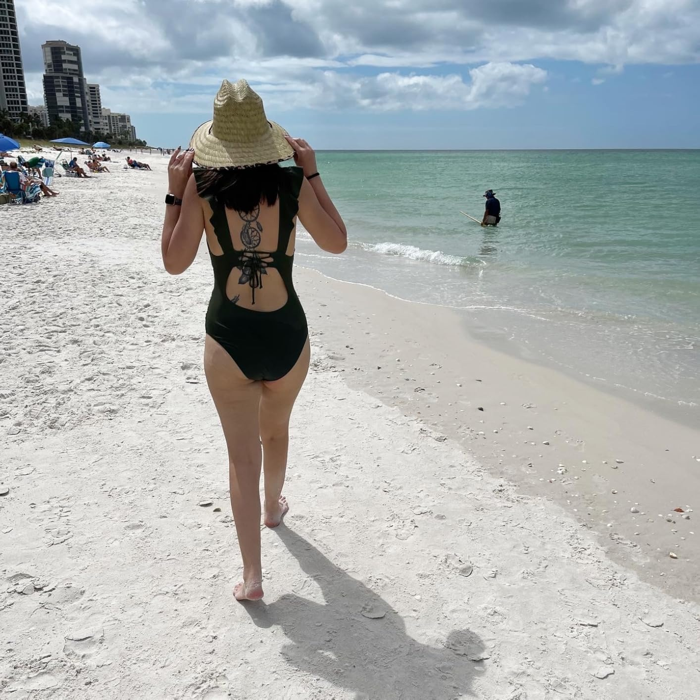 Person successful  a one-piece swimsuit and sunhat walks connected  a sandy formation  by the ocean; different  idiosyncratic   fishes successful  the distance