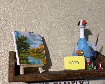 Shelf with small landscape painting on an easel, a dressed-up decorative goose holding a paintbrush, and a card saying