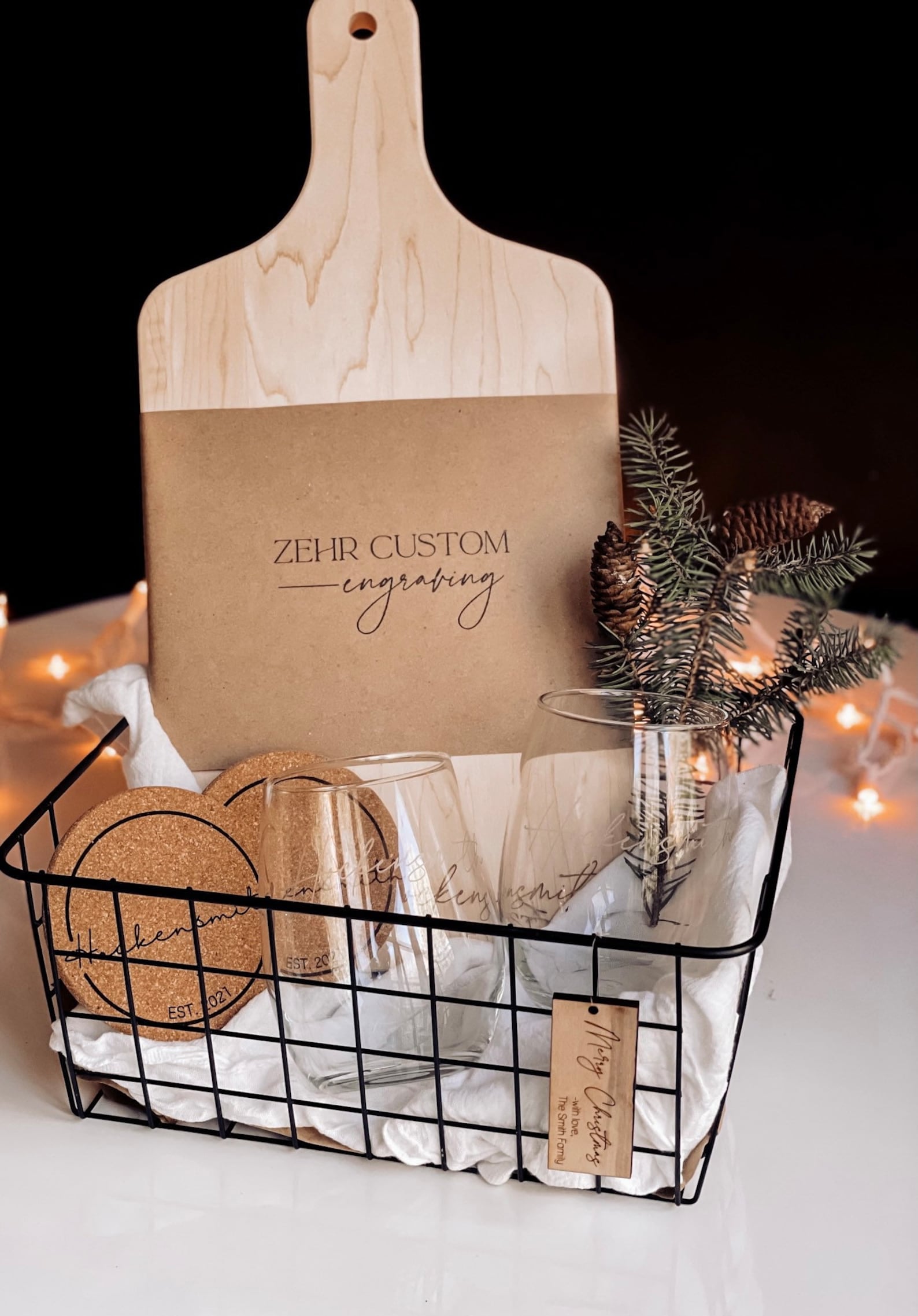 Black basket with engraved wine glasses, engraved cork coasters, and an engraved charcuterie board on top of a white table with twinkle lights