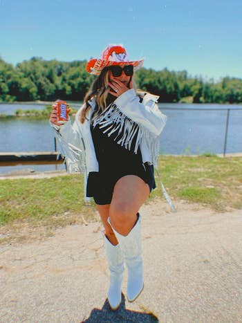 A person posing with a beverage by a lake, wearing a fringed jacket, hat, sunglasses, and boots