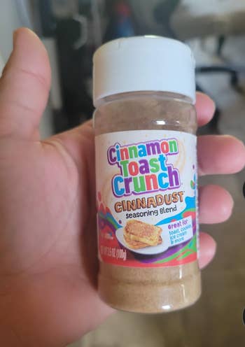Hand holding a vessel  of Cinnamon Toast Crunch Cinnadust seasoning blend, labeled arsenic  large  for toast, cookies, crystal  cream, and more