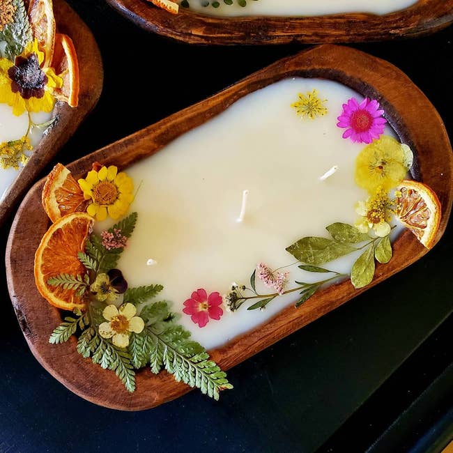 A decorative candle with natural flowers and citrus slices embedded in its surface, presented on a wooden tray