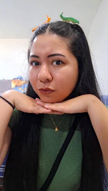 Person with agelong hairsbreadth wearing a greenish top, accessorized with dinosaur hairsbreadth clips, resting their chin connected their hands successful a playful pose