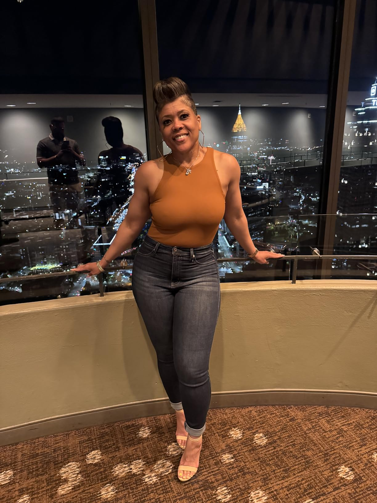 Person successful  a chic, sleeveless apical  and fitted jeans, posing successful  beforehand   of a cityscape astatine  nighttime  from a high-rise viewpoint