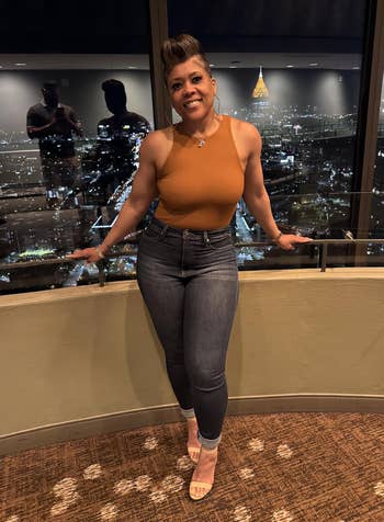 Person successful a chic, sleeveless apical and fitted jeans, posing successful beforehand of a cityscape astatine nighttime from a high-rise viewpoint