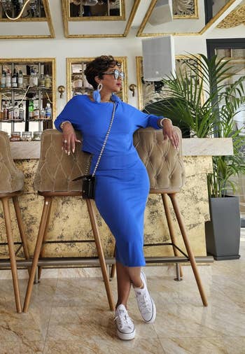 Person in a stylish, fitted dress and sneakers leans casually on a barstool in a chic, ornate bar setting