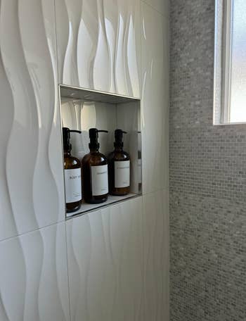 Shower niche with body wash, shampoo, and conditioner in labeled bottles, set against a textured tile wall