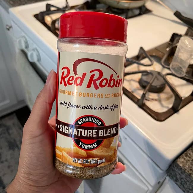 A hand holds a Red Robin Original seasoning blend bottle in a kitchen, near a stove. The label reads