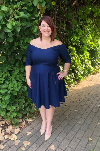 Reviewer wearing knee length flared navy blue off the shoulder dress with three-quarter sleeves and nude heels