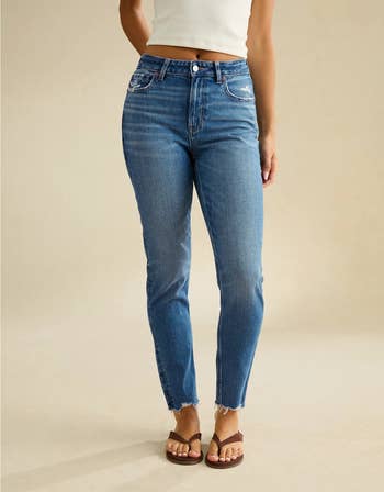 Person modeling fitted jeans with frayed hems, paired with sandals and a sleeveless top. Image for a buying article
