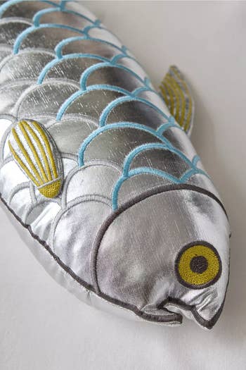 Plush metallic fish-shaped pillow with embroidered scales and fins displayed on a light surface