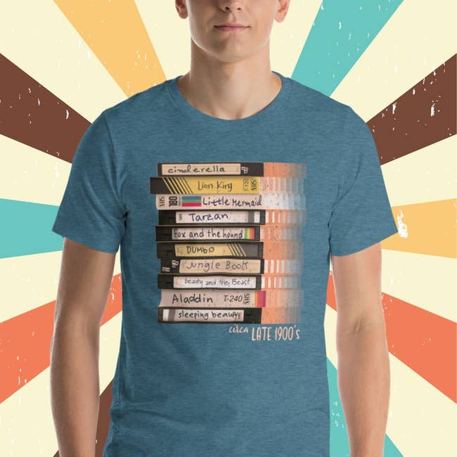 a blue heathered tee with a stack of vhs tapes on it with different disney movie titles handwritten on them