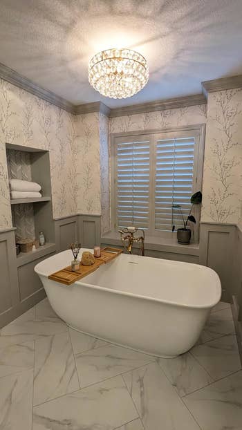 Reviewer's elegant bathroom with a standalone tub with bamboo caddy, chic wallpaper, and a decorative chandelier