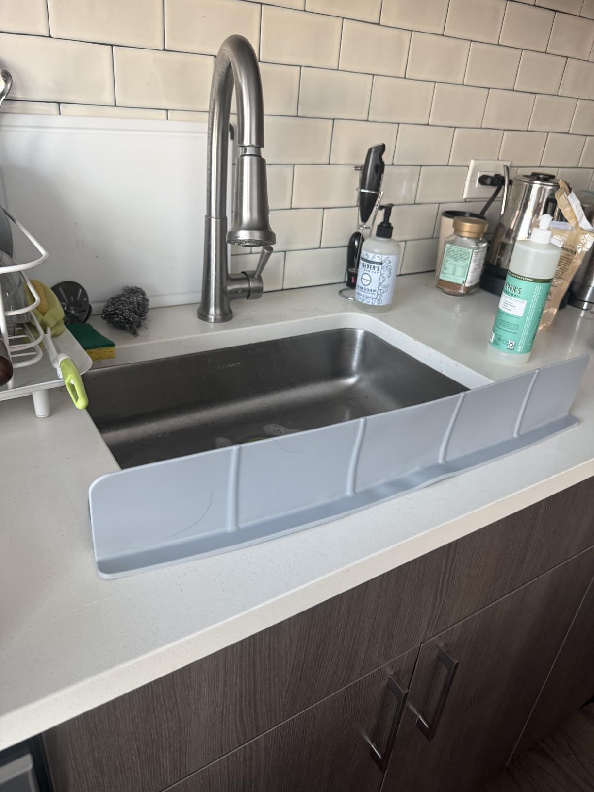 Plastic sink splash guard installed on a kitchen sink with various cleaning supplies nearby