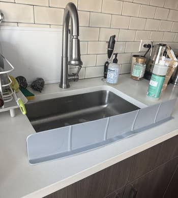 Plastic sink splash guard installed on a kitchen sink with various cleaning supplies nearby