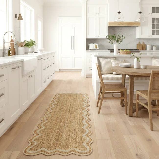 Modern kitchen with white cabinets, a wooden dining table, chairs, and a decorative woven runner on the floor. Natural light fills the space
