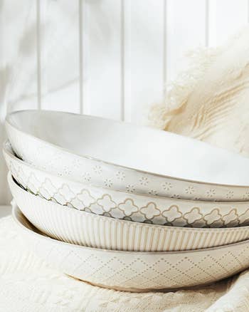 A stack of four decorative white bowls with various patterns on the rims, placed on a light-colored fabric surface