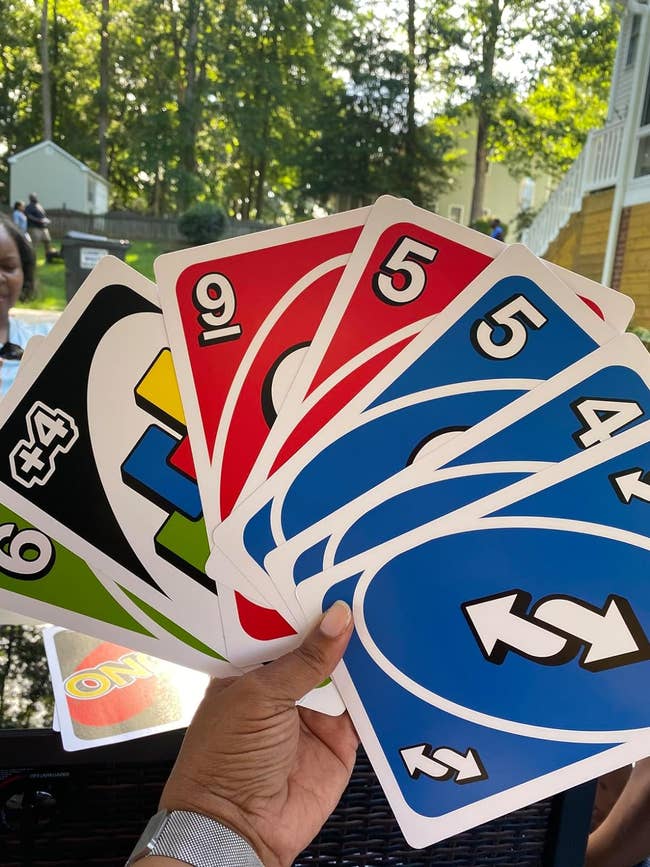 A hand is holding large UNO cards, including a Draw 4 Wild card, a 9, a 5, a 6, and a Reverse, all from different color sets. Trees and houses are visible in the background