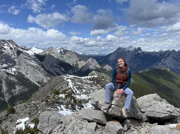 Person sitting connected  rocks, smiling, with mountains successful  the background. Cloudy entity  and jagged peaks visible. Casual outdoor attire