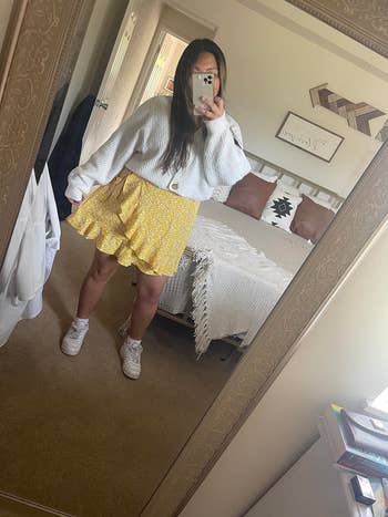 A person takes a mirror selfie wearing a textured top, a patterned skirt, and sneakers. Bedroom decor visible in the background