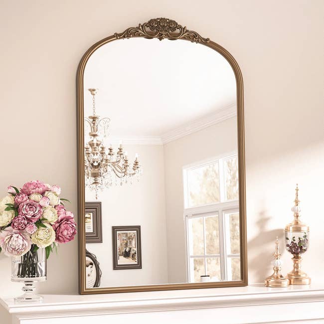 Elegant mirror with ornate frame on mantel, reflecting a chandelier, window, and artwork; vase of flowers and decorative items beside it