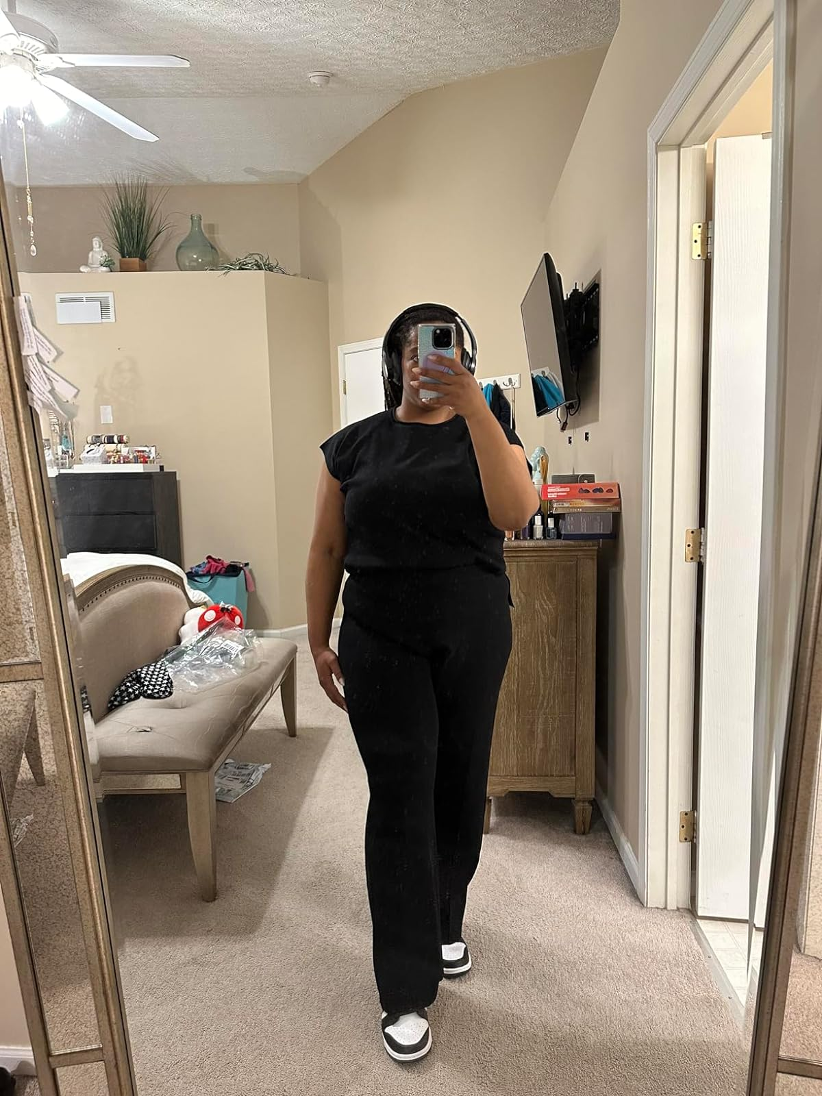 Person taking mirror selfie wearing a black top and pants with sneakers. A cozy room setting is reflected in the background
