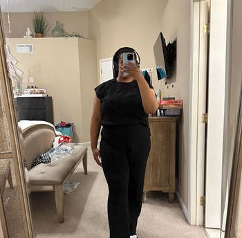 Person taking mirror selfie wearing a black top and pants with sneakers. A cozy room setting is reflected in the background
