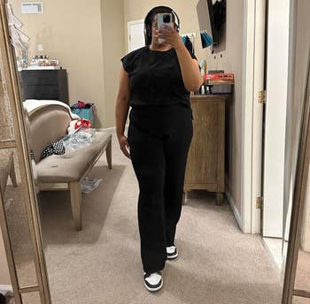 Person taking mirror selfie wearing a black top and pants with sneakers. A cozy room setting is reflected in the background