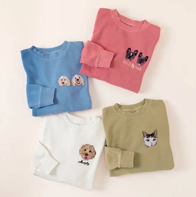 Sweaters in blue, coral, white, and sage green that all have pets embroidered on the upper left chest 