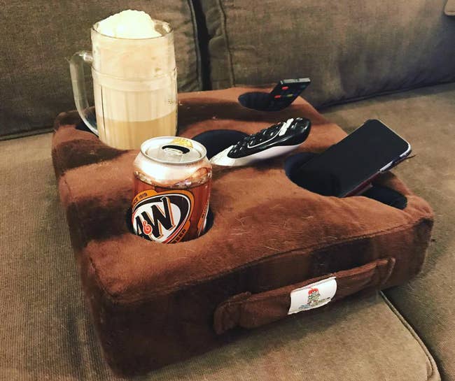 A brown pillow with five holes fitting a can, a drink, a remote control, and a phone 