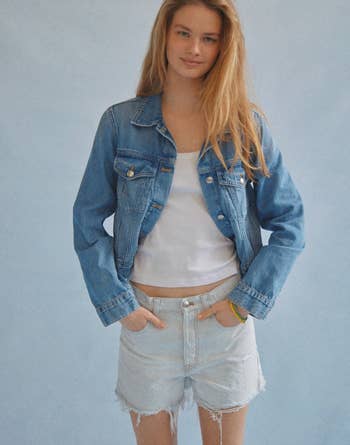 Model in a denim jacket and frayed denim shorts with hands in pockets, posing casually against a plain background