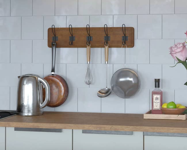 Wooden five hook rack installed on a kitchen wall holding kitchen utensils and pans 