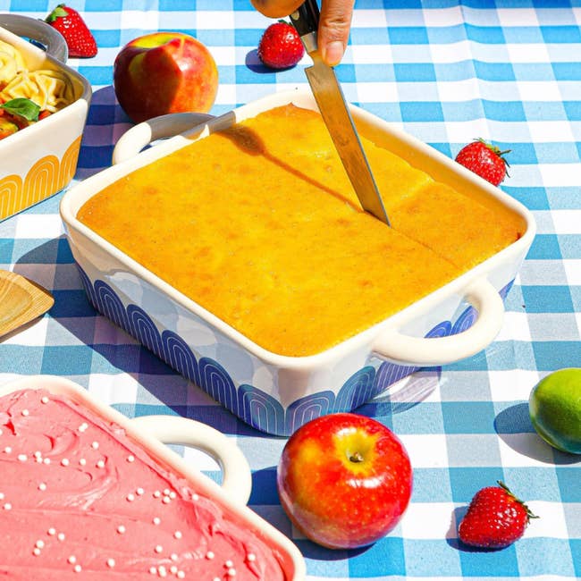 A crockery of cornbread is being sliced connected a checkered tablecloth, surrounded by apples and strawberries