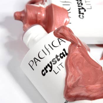 Two bottles of Pacifica Crystal Lit highlighter with iridescent liquid spilled implicit    them, highlighting the product's shimmer and appeal