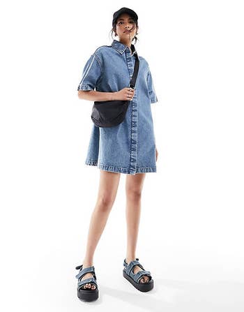 Model in denim dress with black sandals, cap, and a crossbody bag, posing for a shopping ad