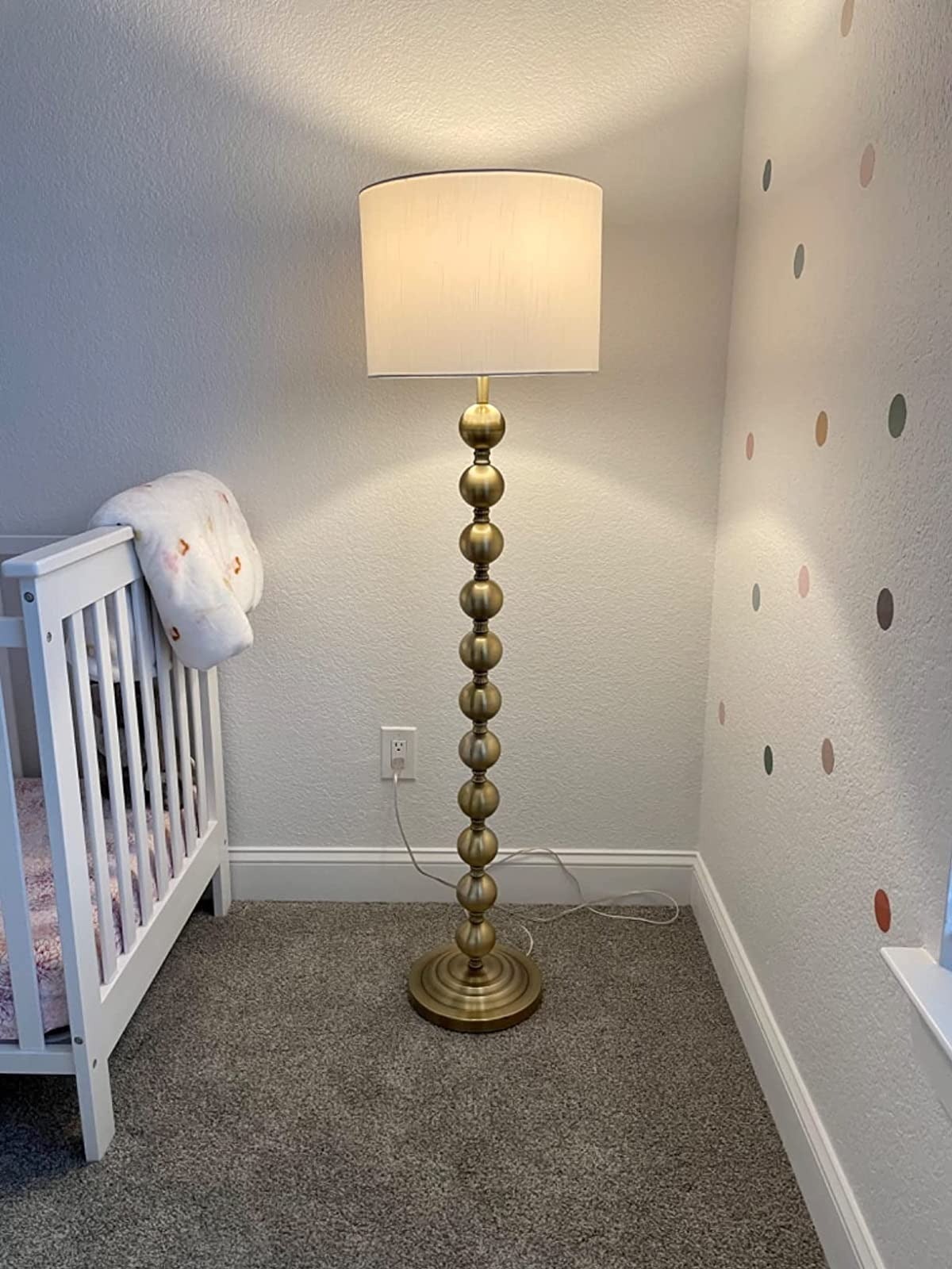 Reviewer image of gold curved modern floor lamp with with lampshade plugged into corner of nursery next to a white crib and polka dot wall