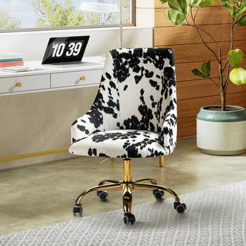 Stylish office chair with a black and white cow print design on a plush seat, set on adjustable gold legs with wheels, in a modern workspace