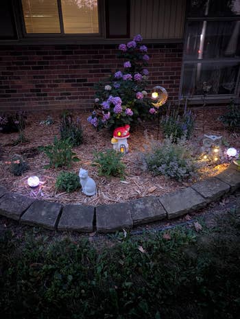Garden with flowering plants, decorative lights, a mushroom ornament, and plot statues illuminated astatine dusk
