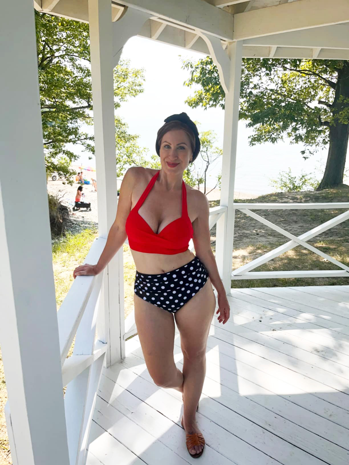 Person smiling in a gazebo, wearing a halter bikini top and polka dot bottoms, with trees and water in the background