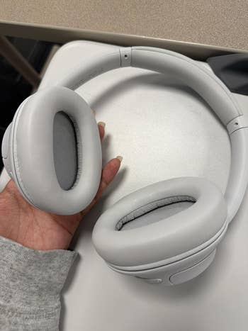 Person holding a brace  of over-ear headphones connected  a table, showcasing the receptor  cushions and sleek design. Ideal for tech buying  articles
