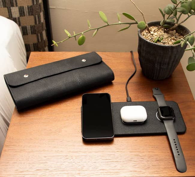a leather folded case sitting next to the charging pad in black with a phone, smart watch, and wireless earbuds charging on it 