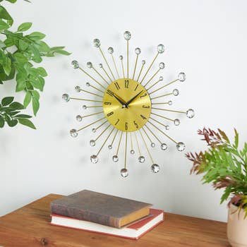 A decorative wall clock with a sunburst design, featuring crystal accents, hangs above a wooden table with stacked books and plants nearby