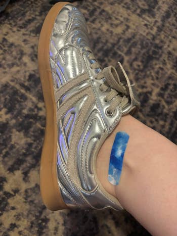 A metallic sneaker-wearing foot with a blue bandage on the ankle. The background is a textured carpet
